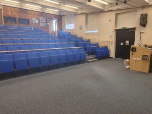 An image of inside a lecture theatre on Highfield Campus, University of Southampton. The picture shows rows of blue tiered seating. The picture is taken from the front of the room where the lecturer would stand.
